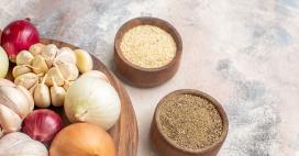 A wooden board holds a collection of culinary delights: red and white onions, fragrant garlic bulbs, and two wooden bowls filled with spices. The photo is shot in bright daylight, with a focus on the natural textures and colors of the ingredients, creating an inviting and wholesome mood. 