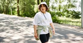  A woman with wavy dark hair walks on a paved path flanked by a verdant landscape. She is wearing a white t-shirt and black shorts, with a green sweater tied around her waist and a white hat. The background shows trees and green vegetation.    