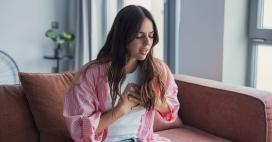 A young woman with wavy brown hair sits on a couch in a home interior. She wears a pink striped shirt, white t-shirt, and light blue jeans. She clutches her chest with both hands and appears to be in distress. The scene is brightly lit by daylight streaming in from nearby windows