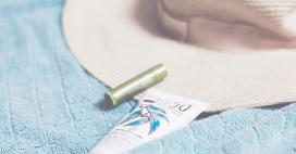 A tube of sunscreen with a leafy design, a green lip balm, and a wide-brimmed straw hat are neatly arranged on a textured, light blue towel, evoking thoughts of warmth and outdoor activities. 