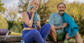 A woman with white hair and a man with a beard relax on stone steps outdoors after exercising. The woman wears a blue top and leggings and dries her face with a blue towel. The man wears a light blue long-sleeved shirt with green shorts, and a blue towel is draped over his shoulder.