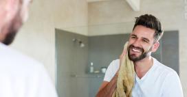 A happy man with dark hair and a beard stands in front of a mirror, drying his face with a tan towel in a bright bathroom. The image has a clean and fresh feel, suggesting grooming, wellness, and personal care