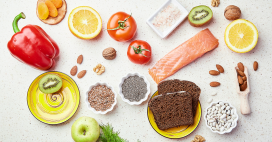 Overhead shot of healthy fruits, vegetables, nuts, seeds and grains arranged on a white surface. A variety of colorful produce creates an appealing flat lay.