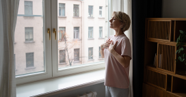 A mature woman with short blonde hair stands in a softly lit room near a large window, hands gently placed on her chest. She wears a pink t-shirt and grey pants.