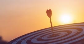 A dart hits the center of a round dartboard at sunset, creating a warm and dreamy feeling. The dartboard is in focus, but the sun and clouds are beautifully blurred in the background, creating an abstract look.