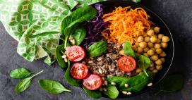 A vibrant vegan bowl is pictured from directly above, containing an assortment of fresh vegetables and grains. There is shredded orange carrots, red cabbage, chickpeas, spinach, tomatoes, grains and pine nuts in a black bowl.