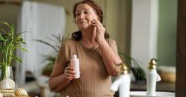 A senior woman with brown hair stands in a bathroom, holding a bottle of pink lotion. She is in front of a mirror, and has applied lotion to her face. The bathroom has neutral decor, with a white sink and leafy green plants. 