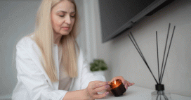 An adult woman with long blonde hair is lighting a small, amber-colored candle. The setting is indoors, possibly a home, with a white surface holding the candle and an aroma diffuser with black reeds. Above is a dark, flat screen television mounted on the wall. The soft lighting contributes to a calm mood.