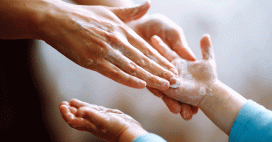 A close-up shot captures an adult carefully applying soap onto a child's hands. The hands are wet and covered in white soap.