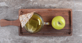A top-down, bright shot depicts a clear mug of tea with a tea bag steeping, a triangular rice cake, and a whole green apple on a dark brown wooden tray. The tray sits on a grey and white mottled surface.