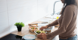 A woman wearing a brown sweater and blue jeans stands in a bright, modern kitchen, organizing fresh meals into clear glass storage containers. The wooden countertop holds several prepared dishes, including a vibrant green salad topped with sliced avocado and a pasta salad with cherry tomatoes and herbs. 